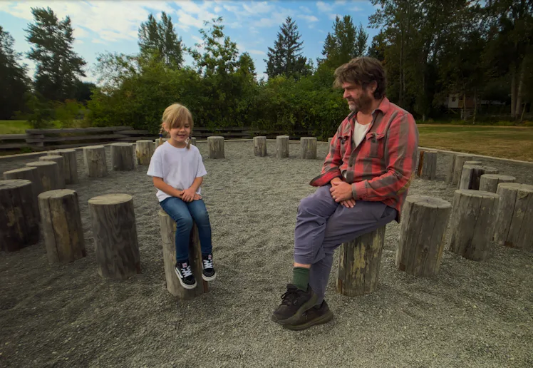 Zach Galifianakis sits on a tree stump talking to a child in a screen still from This Is A Gardening...