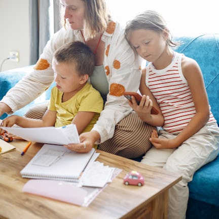 A person sits on a blue sofa with two children, looking at documents and a tablet. One child holds a...