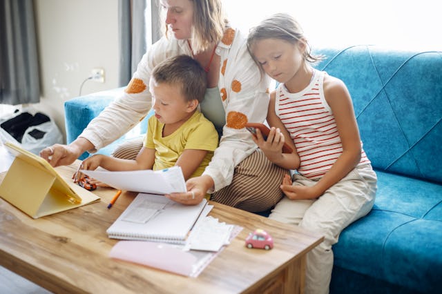 A person sits on a blue sofa with two children, looking at documents and a tablet. One child holds a...