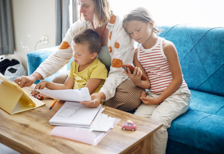 A person sits on a blue sofa with two children, looking at documents and a tablet. One child holds a...
