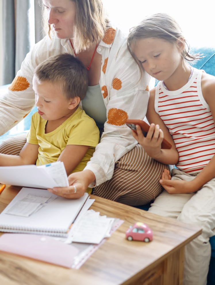 A person sits on a blue sofa with two children, looking at documents and a tablet. One child holds a...