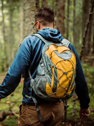 Two people with backpacks hike through a dense, green forest. One is in focus, wearing a blue jacket...