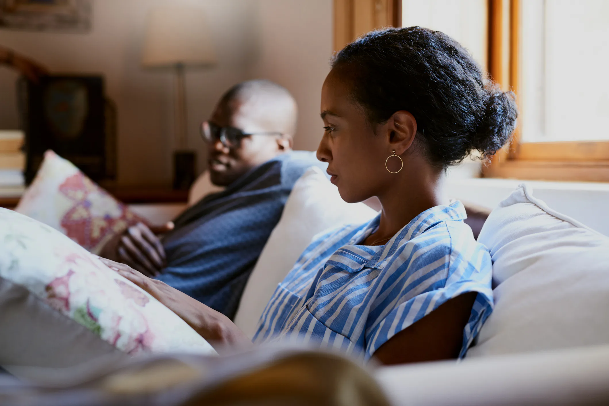 Two people sit on a couch indoors. The person in the foreground wears a blue striped shirt and earri...