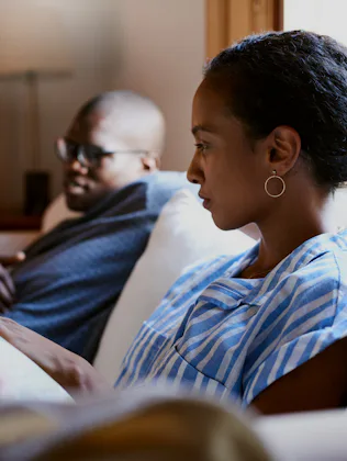 Two people sit on a couch indoors. The person in the foreground wears a blue striped shirt and earri...