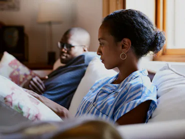 Two people sit on a couch indoors. The person in the foreground wears a blue striped shirt and earri...
