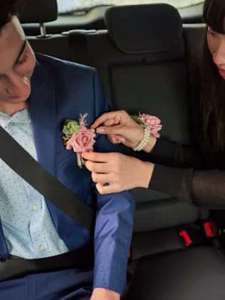 A woman in a car attaches a boutonniere to a man's blue suit. Both are seated and wear seat belts; a...