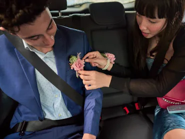 A woman in a car attaches a boutonniere to a man's blue suit. Both are seated and wear seat belts; a...