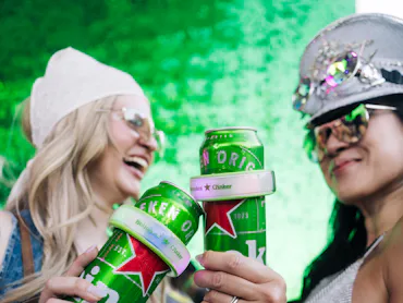 Two people celebrate with canned drinks in hand, dressed in casual festival attire with accessories,...