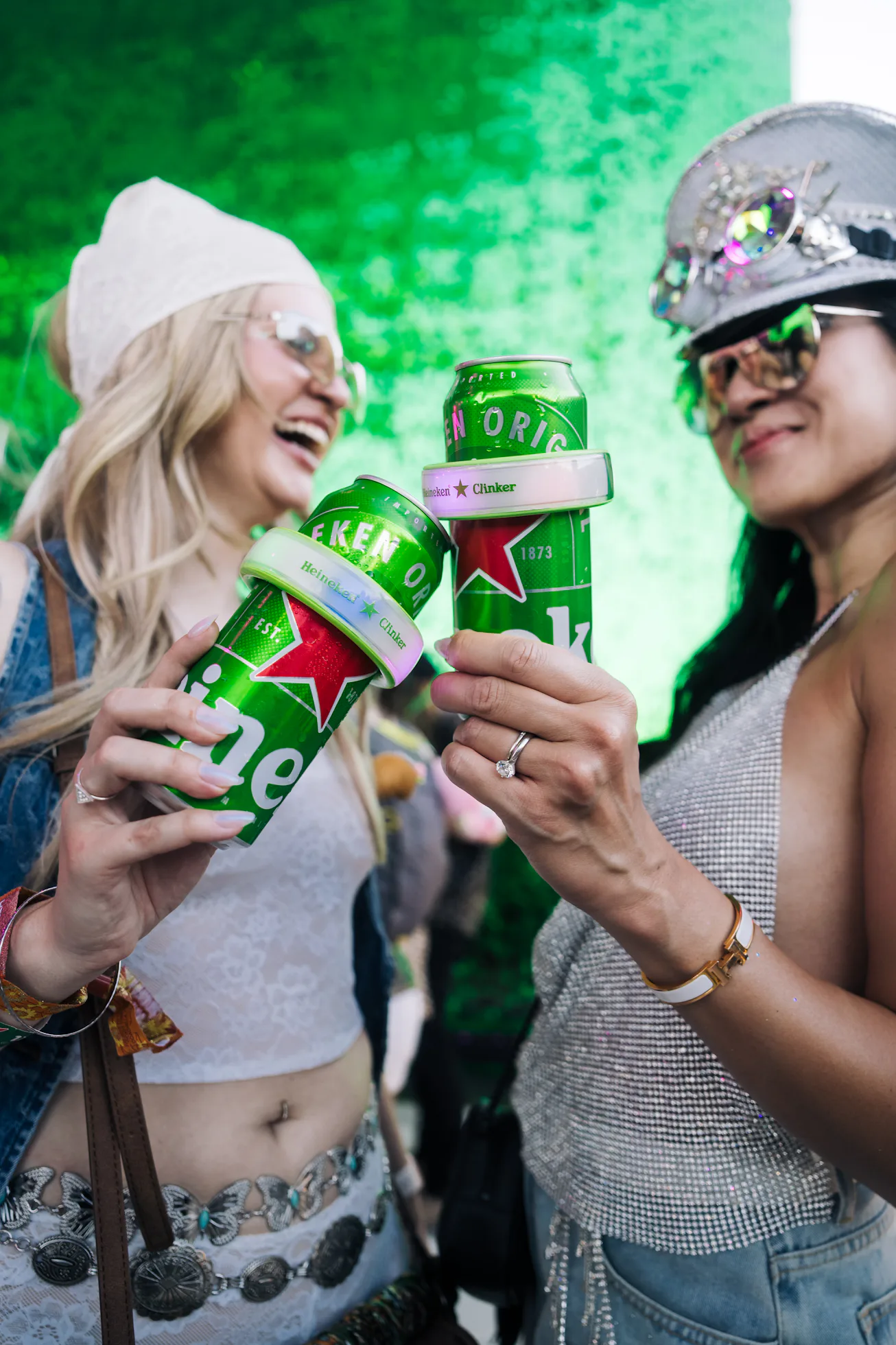 Two people celebrate with canned drinks in hand, dressed in casual festival attire with accessories,...