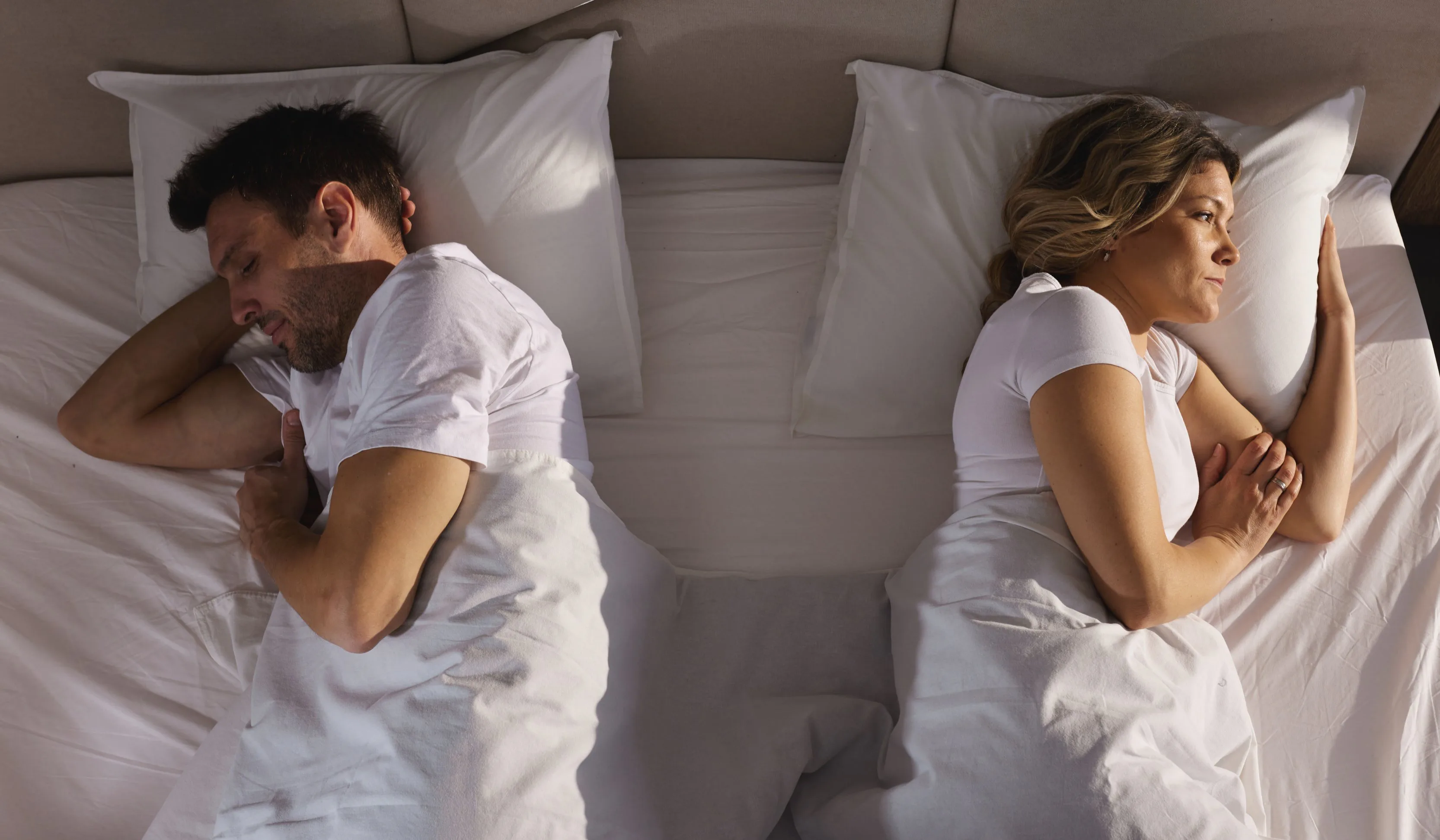 A man and woman lie in bed back-to-back under white sheets, appearing upset or distant, each looking...