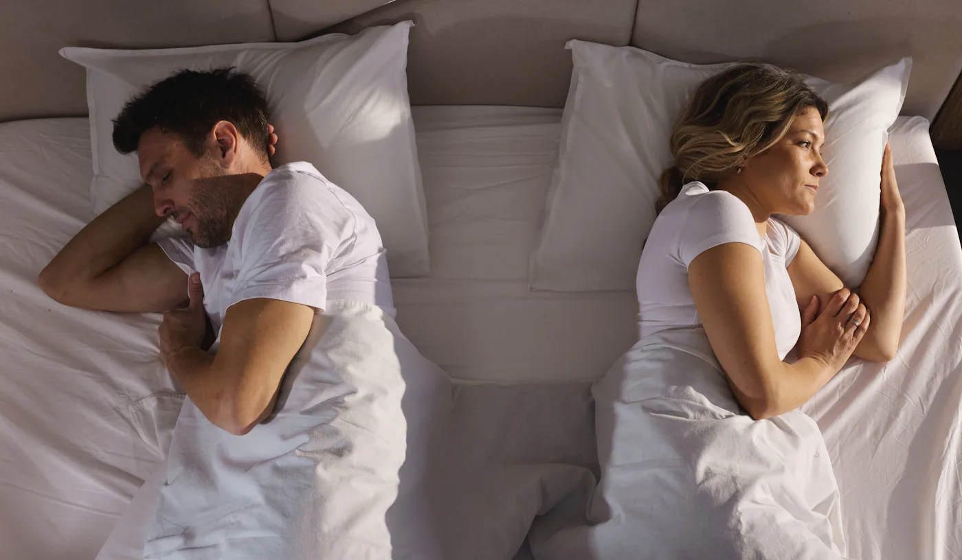 A man and woman lie in bed back-to-back under white sheets, appearing upset or distant, each looking away in opposite directions.