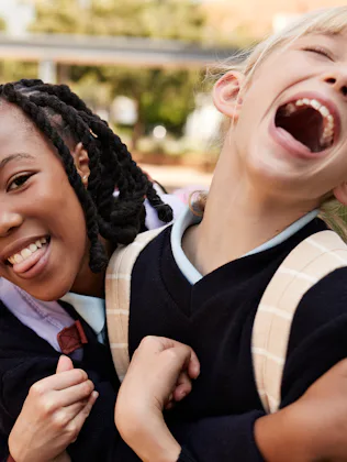 Two young children wearing dark uniforms and backpacks share a joyful moment, one smiling and the ot...