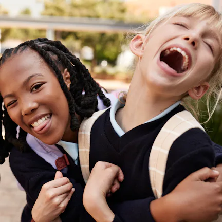 Two young children wearing dark uniforms and backpacks share a joyful moment, one smiling and the ot...