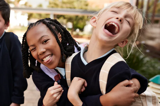 Two young children wearing dark uniforms and backpacks share a joyful moment, one smiling and the ot...