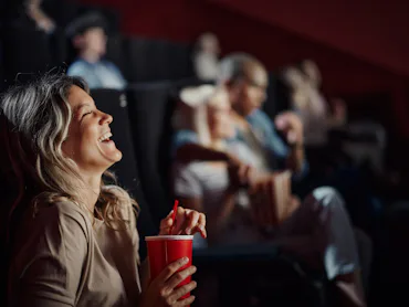 A woman in a cinema laughs while holding a red cup. Other people are seated behind her, some watchin...