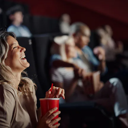A woman in a cinema laughs while holding a red cup. Other people are seated behind her, some watchin...