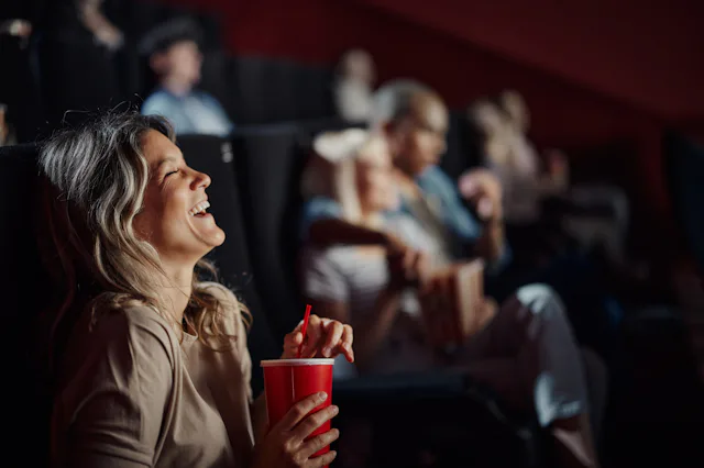 A woman in a cinema laughs while holding a red cup. Other people are seated behind her, some watchin...