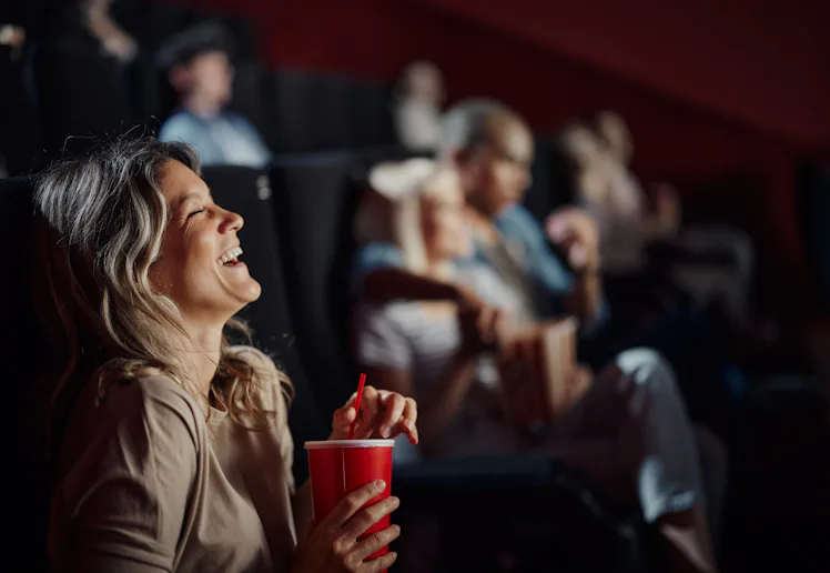 A woman in a cinema laughs while holding a red cup. Other people are seated behind her, some watchin...