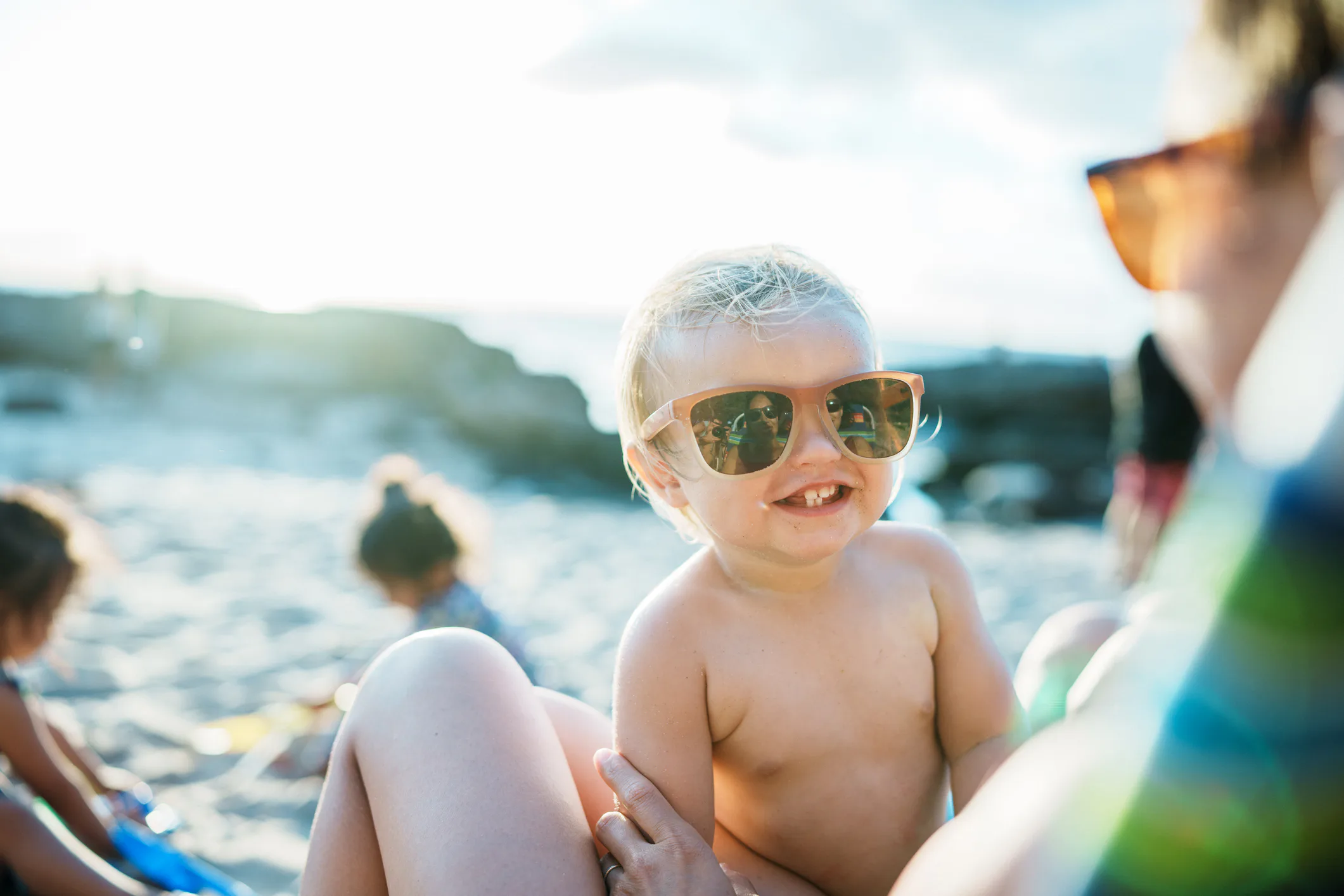 A smiling toddler wearing sunglasses is being held by an adult at a sunny beach. Other children are ...