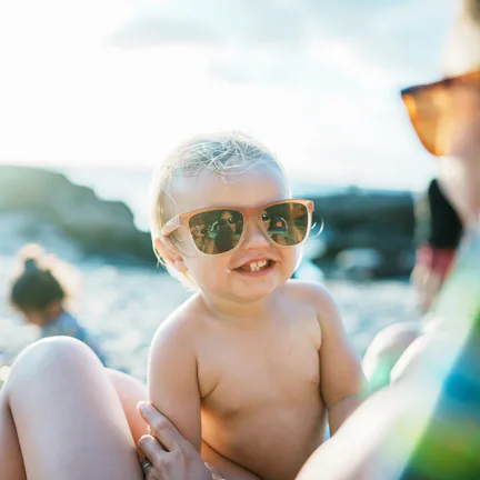 A smiling toddler wearing sunglasses is being held by an adult at a sunny beach. Other children are ...