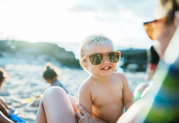 A smiling toddler wearing sunglasses is being held by an adult at a sunny beach. Other children are ...