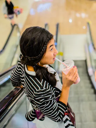 Two people are going up an escalator in a mall, each holding and drinking from a clear cup with a st...