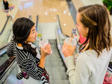Two people are going up an escalator in a mall, each holding and drinking from a clear cup with a st...