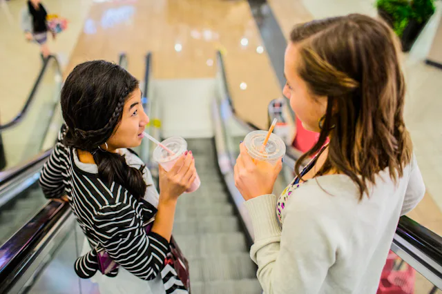 Two people are going up an escalator in a mall, each holding and drinking from a clear cup with a st...