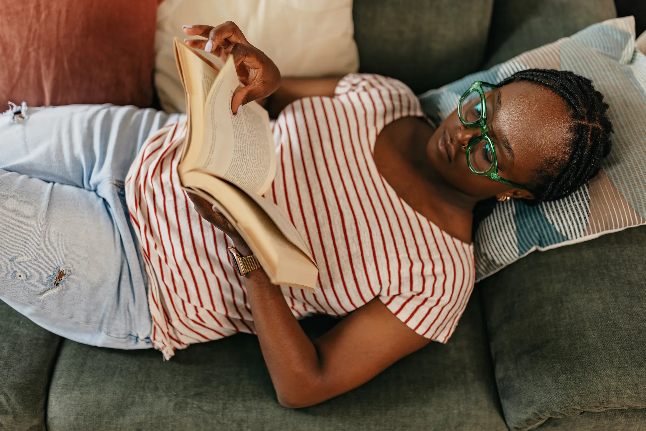 A person with glasses and braids is lying on a sofa, wearing a striped shirt and jeans, reading a bo...