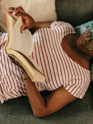 A person with glasses and braids is lying on a sofa, wearing a striped shirt and jeans, reading a bo...