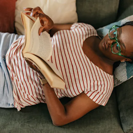 A person with glasses and braids is lying on a sofa, wearing a striped shirt and jeans, reading a bo...
