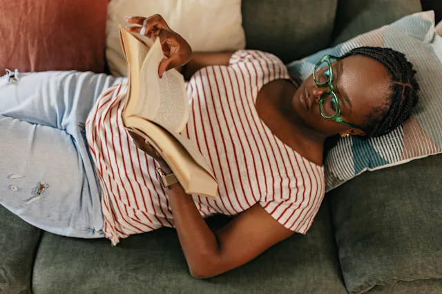 A person with glasses and braids is lying on a sofa, wearing a striped shirt and jeans, reading a bo...