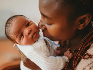 A person gently cradles a baby, giving a tender kiss on the forehead. The baby is wrapped in a white...