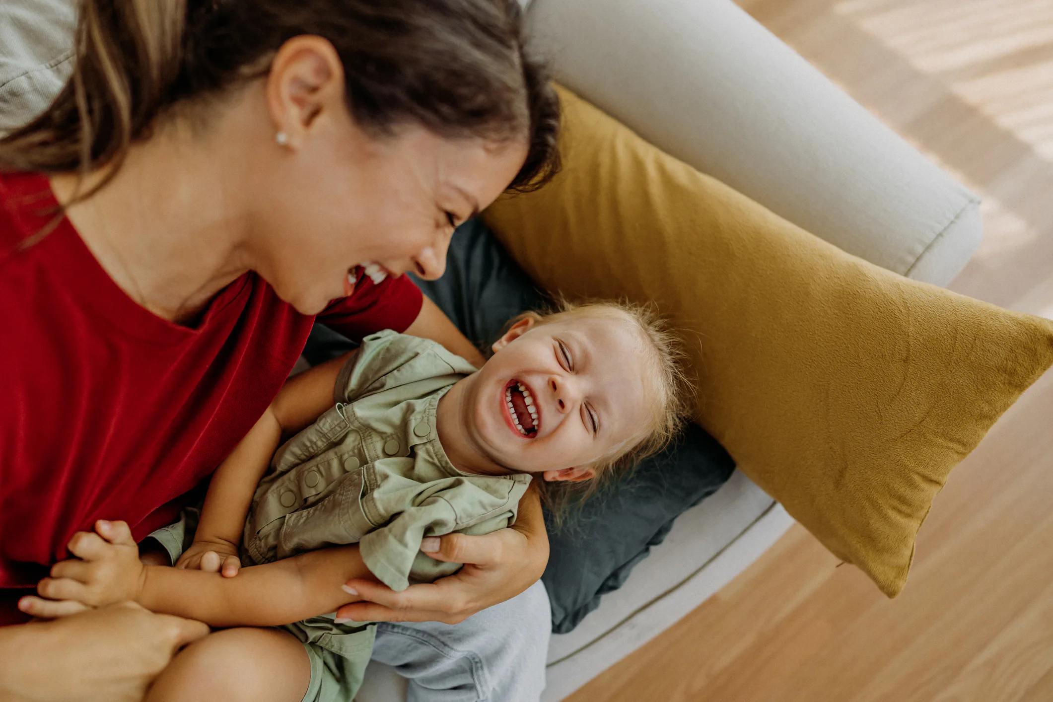 A woman in a red shirt sits on a couch, holding a laughing child in a green dress. They are both smi...