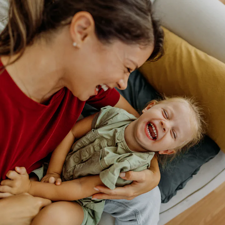 A woman in a red shirt sits on a couch, holding a laughing child in a green dress. They are both smi...