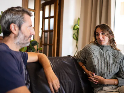 A man and woman are sitting on a couch in a living room. The man is speaking while the woman listens...