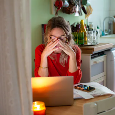 Woman in a kitchen sits at a table with a laptop and phone, rubbing her eyes amid papers and a lit candle, looking tired or stressed.