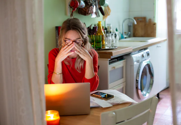 Woman in a kitchen sits at a table with a laptop and phone, rubbing her eyes amid papers and a lit c...