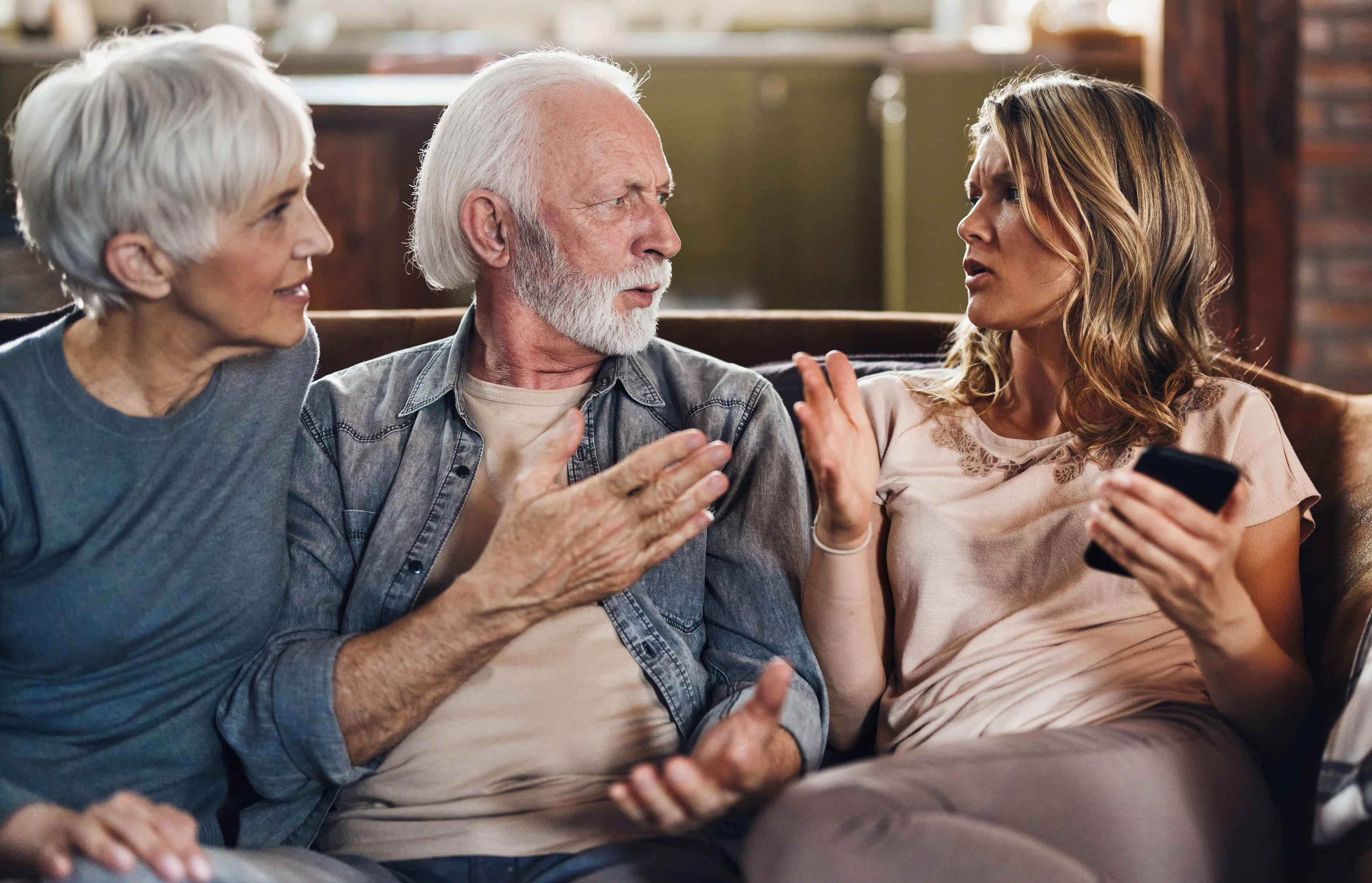 Three adults on a couch: an older white couple arguing with a younger woman holding a smartphone, an...