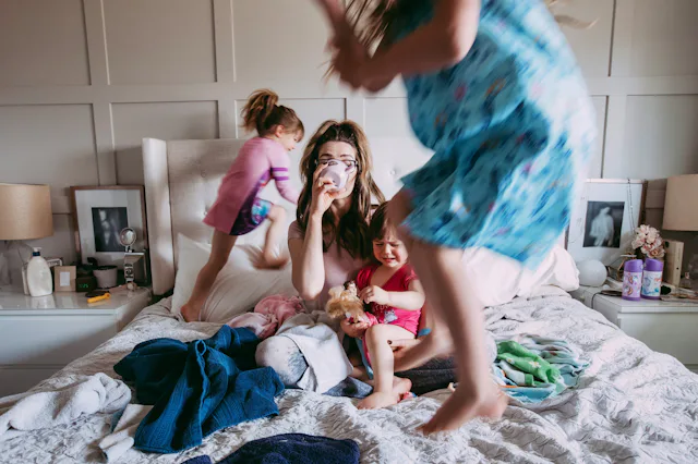 A tired mother sits on a messy bed drinking coffee while three young children play around her; one j...