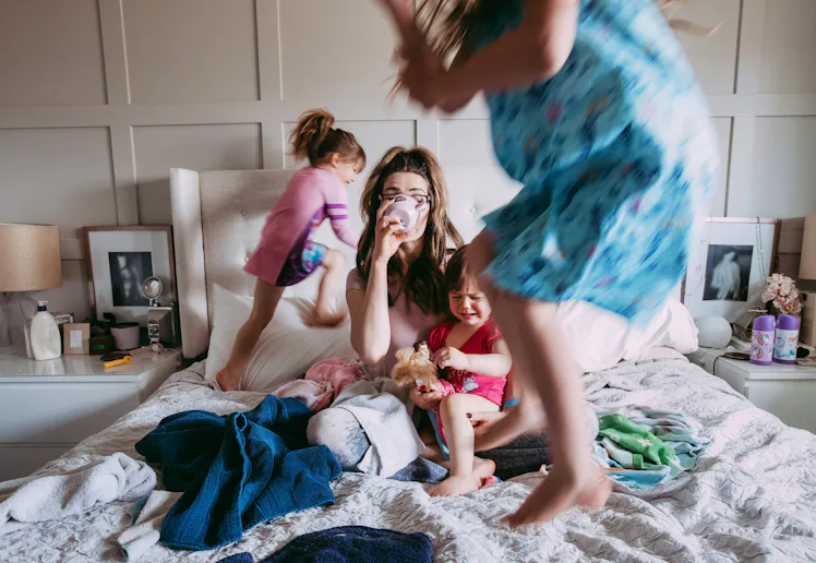 A tired mother sits on a messy bed drinking coffee while three young children play around her; one j...
