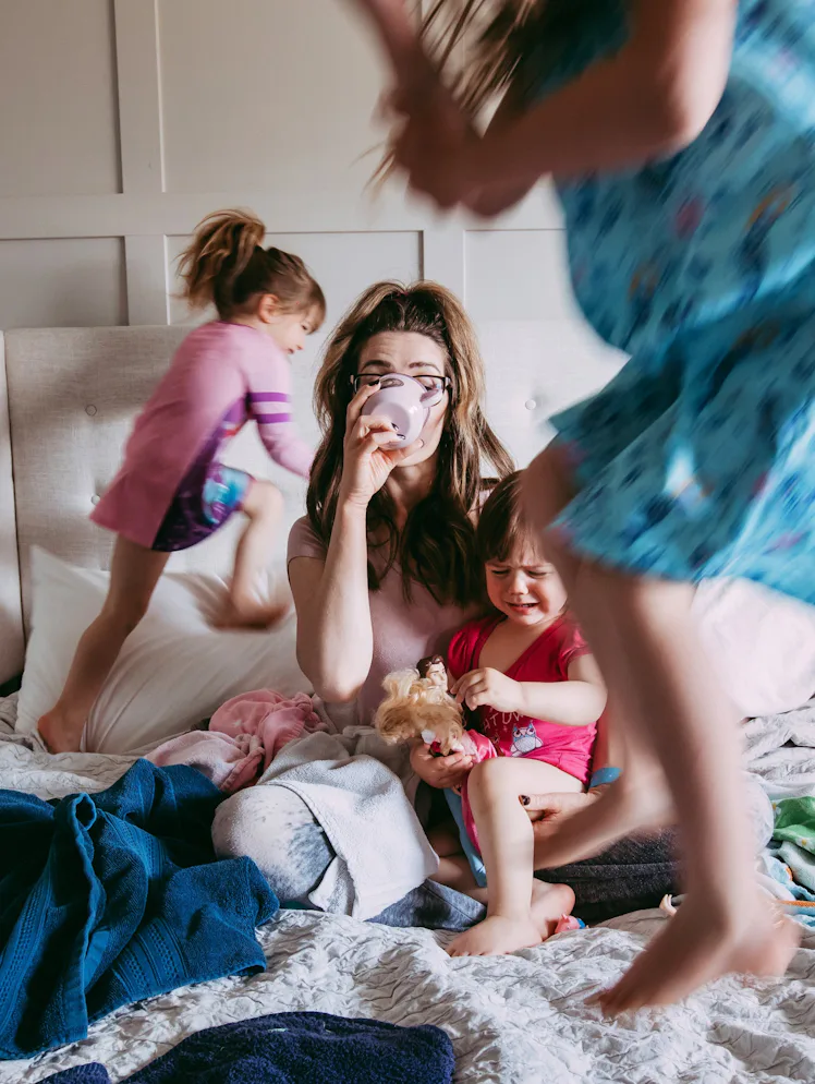 A tired mother sits on a messy bed drinking coffee while three young children play around her; one j...