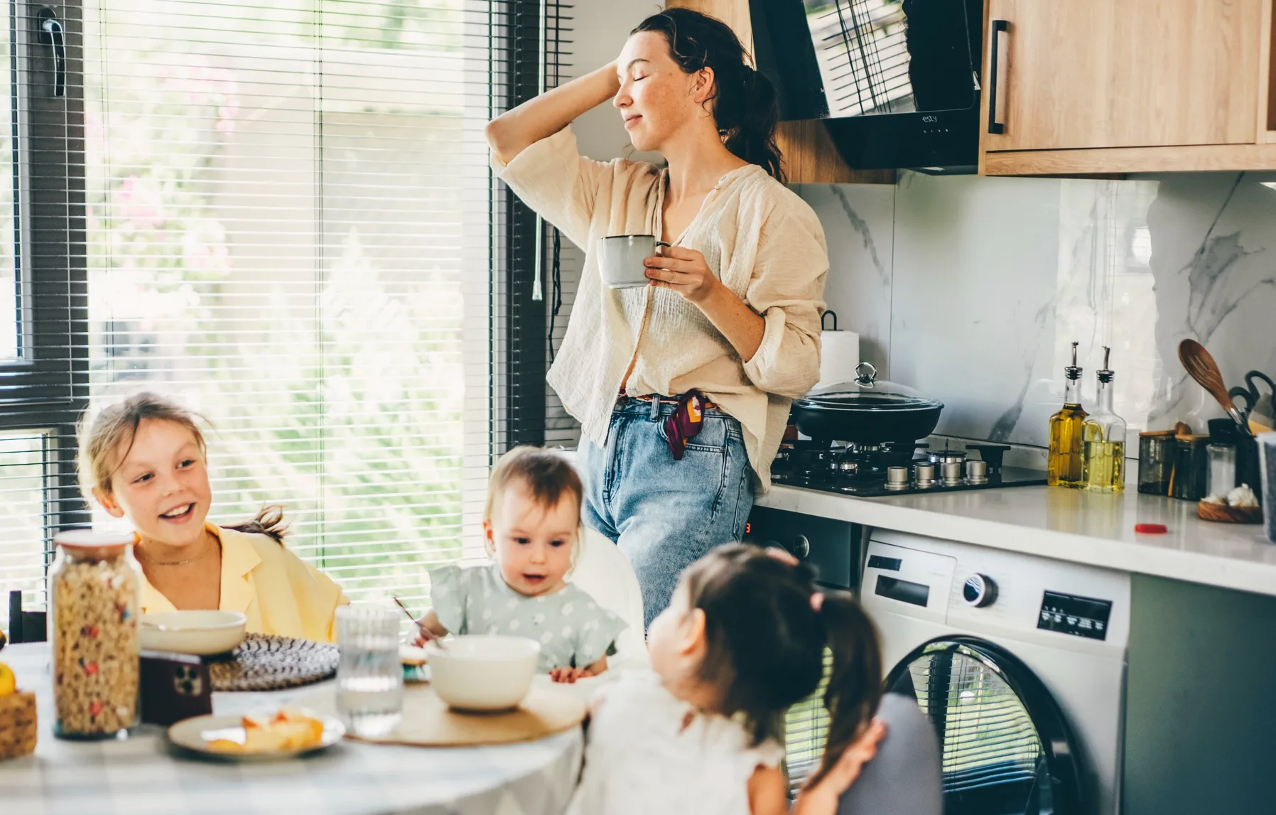 A relaxed mother stands in a sunlit kitchen holding a mug, smiling as three young children sit aroun...
