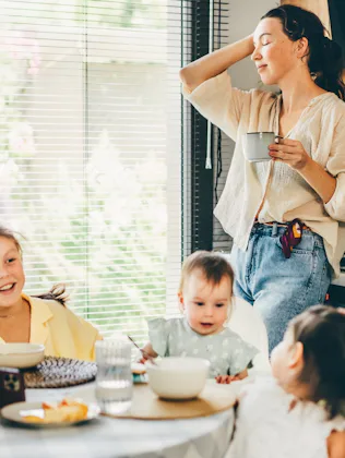 A relaxed mother stands in a sunlit kitchen holding a mug, smiling as three young children sit aroun...