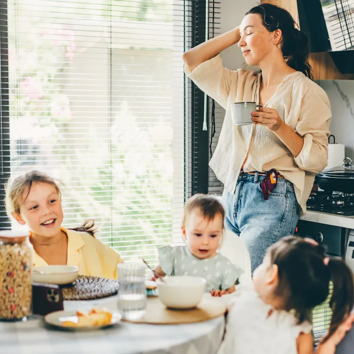 A relaxed mother stands in a sunlit kitchen holding a mug, smiling as three young children sit aroun...