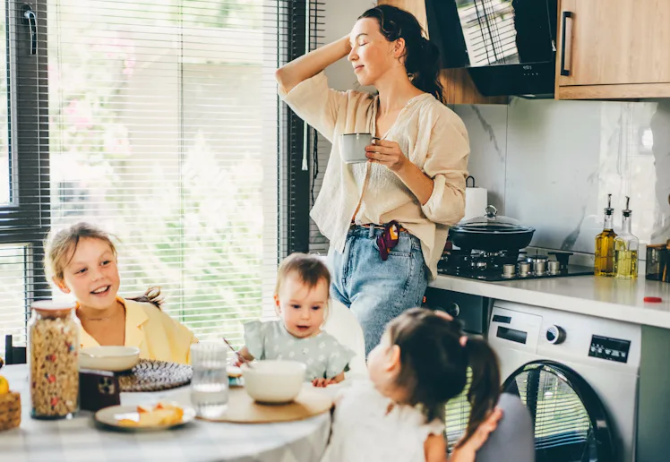 A relaxed mother stands in a sunlit kitchen holding a mug, smiling as three young children sit aroun...