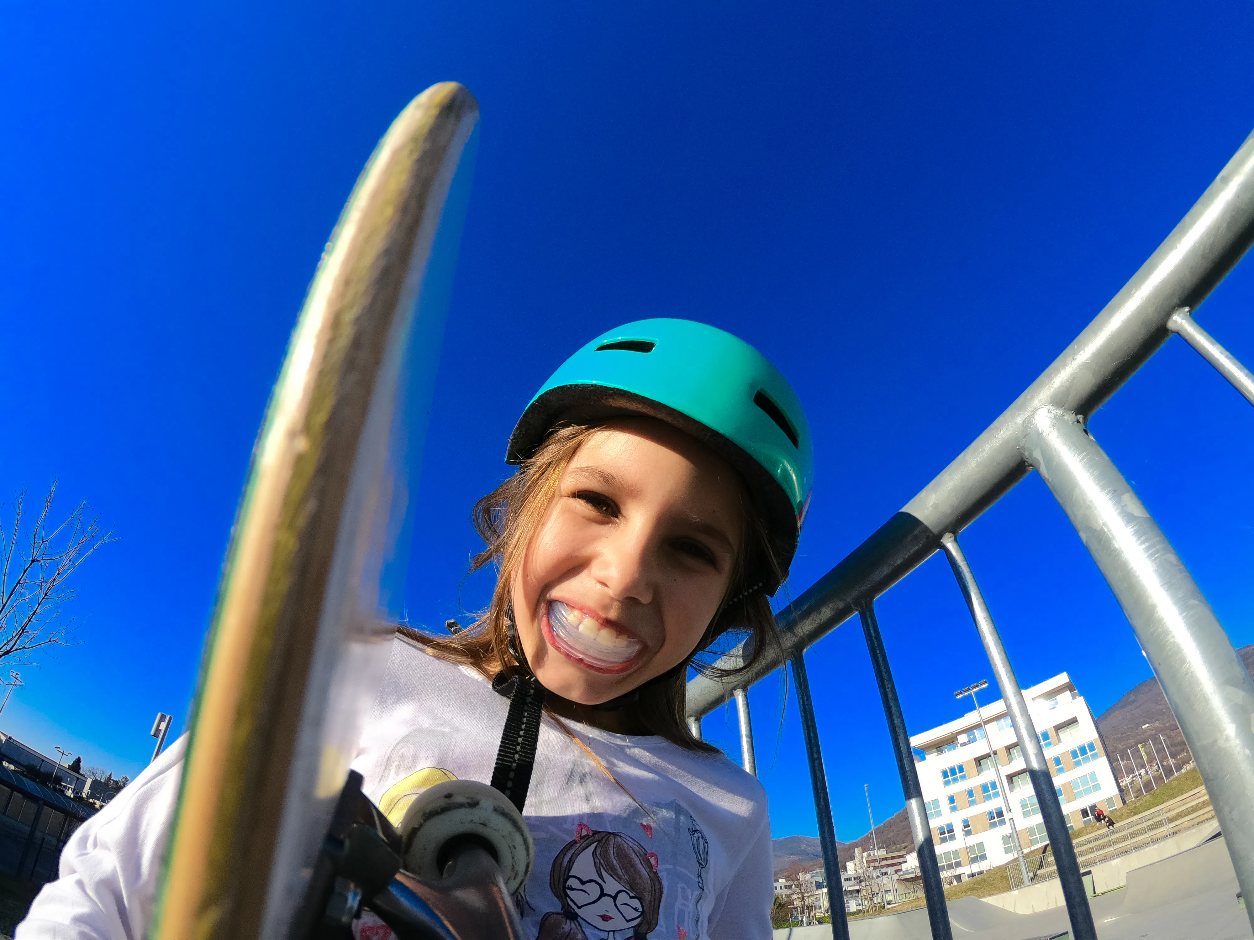 Smiling child wearing a teal helmet holds a skateboard near metal railings at a sunny skate park, br...