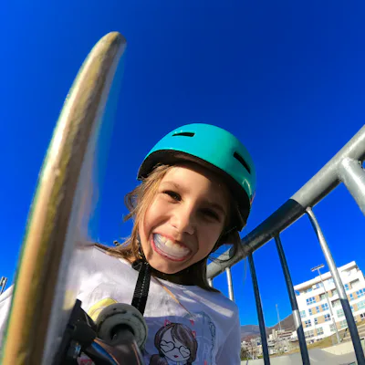 Smiling child wearing a teal helmet holds a skateboard near metal railings at a sunny skate park, bright blue sky and buildings visible in background.