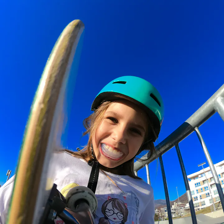 Smiling child wearing a teal helmet holds a skateboard near metal railings at a sunny skate park, br...