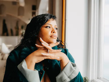 Woman with dark hair and coat resting chin on folded hands, gazing out a window in a softly lit inte...